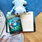 Ein offenes Buch, darauf liegt "The Immortals: Dawn of the Axolotl" von Kit Brooks. Ein blaues Plüschtier daneben.