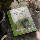 Auf einem Tisch liegt ein Buch mit dem Titel "The Container Garden Book". Daneben Pflanzen und Erde verstreut.
