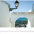 Text: "Arcos de la Frontera". Ansicht einer malerischen, weißen Gasse mit Blick auf Felder und Landschaft durch einen Bogen.