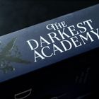 "THE DARKEST ACADEMY II" in weißen Buchstaben auf einem dunkelblauen Buchrücken. Verzierungen mit einem Raben.