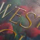 Text in goldener Prägung: "WEST". Im Hintergrund rote Blüten und grüne Blätter, kunstvoll gestaltet.