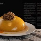 "Gefüllter Käse" steht in der oberen linken Ecke. Eine goldene Käsekugel mit Fleischfüllung liegt auf einem weißen Teller, umgeben von Soße. Der Hintergrund ist schwarz, und die Tischdecke darunter ist spitzenartig gemustert. Links befinden sich detaillierte Beschreibungen des Gerichts.