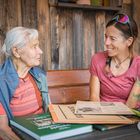 Zwei Frauen sitzen lächelnd auf einer Holzbank. Auf dem Tisch liegen alte Zeitungen und ein grünes Buch. Eine Pflanze ist rechts.