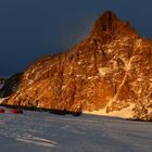 Helle Bergspitze im Abendlicht, zwei rote Zelte unten auf schneebedecktem Boden, vor dunklem Himmel.