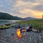Ein Lagerfeuer am Ufer eines Sees, Kajaks und Fahrräder liegen am steinigen Strand, ein Zelt im Hintergrund, Abenddämmerung.