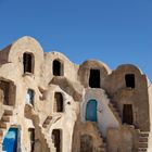 Lehmarchitektur mit Treppen und Bögen vor blauem Himmel. Türen in verschiedenen Farben, einige sind blau gestrichen.