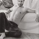 09. Ein Mann sitzt entspannt auf einem Sofa, trägt Jeans, ist oberkörperfrei und blickt lächelnd in die Kamera.