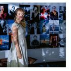 Eine Frau in einem hellen Kleid steht vor einer Fotowand, die mit verschiedenen Porträts und Bildern bedeckt ist.