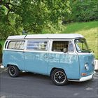 Ein hellblauer, vintage Minibus mit weißem Dach steht am Straßenrand, umgeben von Bäumen und Grasflächen.