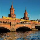 Eine gelbe U-Bahn fährt über die rote Backsteinbrücke, die sich über einen Fluss erstreckt, mit zwei Türmen im Hintergrund.