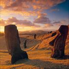 Monumentale Steinstatuen bei Sonnenuntergang auf sanften Hügeln, umgeben von einem dramatischen, orangefarbenen Himmel.