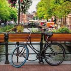 Fahrrad lehnt an Brücke mit blühenden Pflanzen; dahinter ein Kanal mit Booten und am Ufer grüne Bäume.