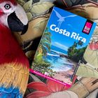 Text: "Costa Rica" und "Reiseführer für individuelles Entdecken". Ein Buch mit Strandfoto auf tropischem Hintergrund. Papageienschnitzerei.
