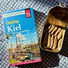 "CityTrip Kiel mit Kieler Förde" auf einem Reiseführer neben einer geöffneten Sardinendose auf einem blau karierten Tuch.