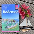 „Bodensee“ und „Reiseführer für individuelles Entdecken“ auf einem blauen Buchcover neben einem kleinen Segelboot und roten Blüten.