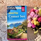 „Mein Trip Comer See“ von Markus Bingel. Reiseführer auf einem Häkeldeckchen, daneben ein bunter Blumenstrauß.