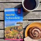 "Mein Trip Schweden" Reiseführer, Holztisch, Tasse Kaffee, Zimtschnecke auf gelbem Teller, illustriertes Papiertuch.