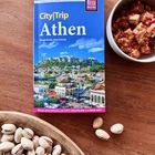 Reiseführer "CityTrip Athen" auf Holztisch, neben Pistazien in Schale und Schüssel mit Käse-Tomaten-Salat.