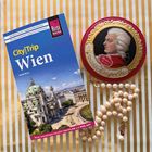 „CityTrip Wien“ Reiseführer, daneben eine Dose Mozartkugeln mit einem Porträt und eine Perlenkette auf gestreiftem Hintergrund.