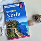 Ein Reiseführer mit der Aufschrift "Insel|Trip Korfu" liegt auf einem Tisch, daneben Muscheln und ein weißes Tuch.