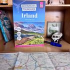 Text: WOHNMOBIL TOURGUIDE Irland mit Nordirland. Illustration eines Reiseführers auf einem Holzregal, umgeben von Deko.