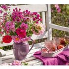 "09" und Kalenderdaten rechts. 
Ein Strauß bunter Blumen in einer lila Vase auf einer weißen Bank. Daneben Obst und Gläser auf einem Tuch.