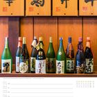 Japanische Schriftzeichen auf Holztafeln, darunter Sake-Flaschen; Kalenderseite mit Februar und Datum 07 markiert.