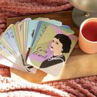 Text: "Coco Chanel." Illustration einer Frau mit Schmuck, Kartenstapel auf Holzbrett, rosa Tasse nebenan auf Decke.