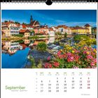 Septemberkalender mit farbenfroher Flussstadt. Blumen im Vordergrund, bunte Gebäude und Kirchturm spiegeln sich im Wasser.