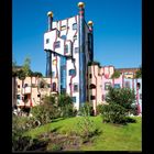 IX 2027. Verspieltes, buntes Gebäude mit unregelmäßigen Formen, goldenen Kugeln und viel Grünfläche im Vordergrund.