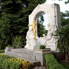 Statue eines goldenen Geigers vor einem weißem Reliefbogen in einem gepflegten, grünen Garten mit Blumen.