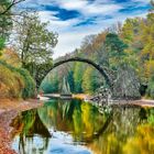 Ein idyllischer Fluss in einem Herbstwald mit einer steinernen, halbkreisförmigen Brücke, die sich im Wasser spiegelt.