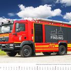 "Freiwillige Feuerwehr Finsing", "FEUERWEHR", "112". Ein rotes Feuerwehrauto steht auf einem Feldweg unter blauem Himmel mit Wolken.