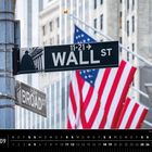 Auf einem Straßenschild steht "Wall St" und "Broad St", im Hintergrund hängen US-Flaggen an Gebäuden.