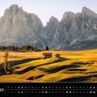 Südtirol 09. Ein Feld mit Holzhütten, sanfte Hügel, Bäume und hohe Berge im Hintergrund in warmem Licht.