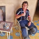 Ein Mann in Motorradkleidung sitzt lächelnd mit einem Glas Tee auf einem Stuhl. Neben ihm steht ein Tisch mit Teekanne.