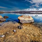Große Felsen liegen im klaren, blauen Wasser eines Sees; im Hintergrund schneebedeckte Berge und blauer Himmel.