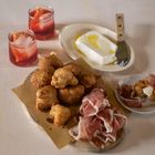 Gebratene Teigbällchen, Prosciutto, Käse mit Öl und zwei rote Getränke auf einem Tisch.