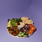 Ein Teller mit Falafel, Pita, frischem Gemüse, Saucenschale, Lemon, Gurken, Zwiebeln und Tomaten. Hintergrund ist lila.