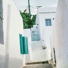 Weiße Gebäude mit türkisfarbenen Fensterläden, eine Katze sitzt auf einer Treppe. Es ist eine ruhige, schmale Gasse.