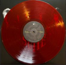 Rote Vinyl-Schallplatte mit mittigem Etikett. Darauf Trackliste und ein kleines weißes Logo. Im Hintergrund verschwommene Silhouetten.