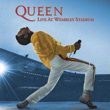 Text: "QUEEN LIVE AT WEMBLEY STADIUM". Eine Person in gelber Jacke und weißer Hose singt dynamisch ins Mikrofon.