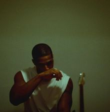 Ein Mann in einem weißen T-Shirt hebt den Arm, verdeckt teilweise sein Gesicht; im Hintergrund steht eine Gitarre.