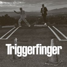 Text: "Triggerfinger". Zwei Männer in Anzügen stehen auf einem Parkplatz, einer zeigt mit der Hand, der andere hält Revers.