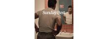 Text "Sunday Best". Ein Mann betrachtet sich im Spiegel eines Badezimmers mit rosa Fliesen und Waschbecken.