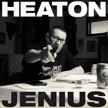 Text: "HEATON" oben, "JENIUS" unten. Ein Mann zeigt mit einem Stift in die Kamera, sitzend an einem Tisch.