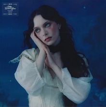 Text: "Holly Humberstone". Frau mit langem, dunklem Haar, weiße Kleidung, Hände an die Wange, blauer Hintergrund.