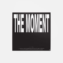 Schwarzer Hintergrund mit großem weißen Text: "THE MOMENT". Darunter kleinere Texte und ein Name.