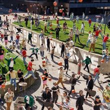 Viele Menschen jonglieren mit Basketbällen, einige tragen Musikinstrumente. Ein lebhafter, bunter Stadtplatz.