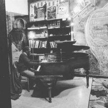 „Mind of Mine“, „The Social Flashflair Company“ an einer Wandtafel. Ein Mensch spielt Klavier in einem Raum voller Bücher.